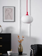 A Clérachic pendant with a red cap and white glass shade beside a framed line drawing, with a black cabinet, vase and chair below.
