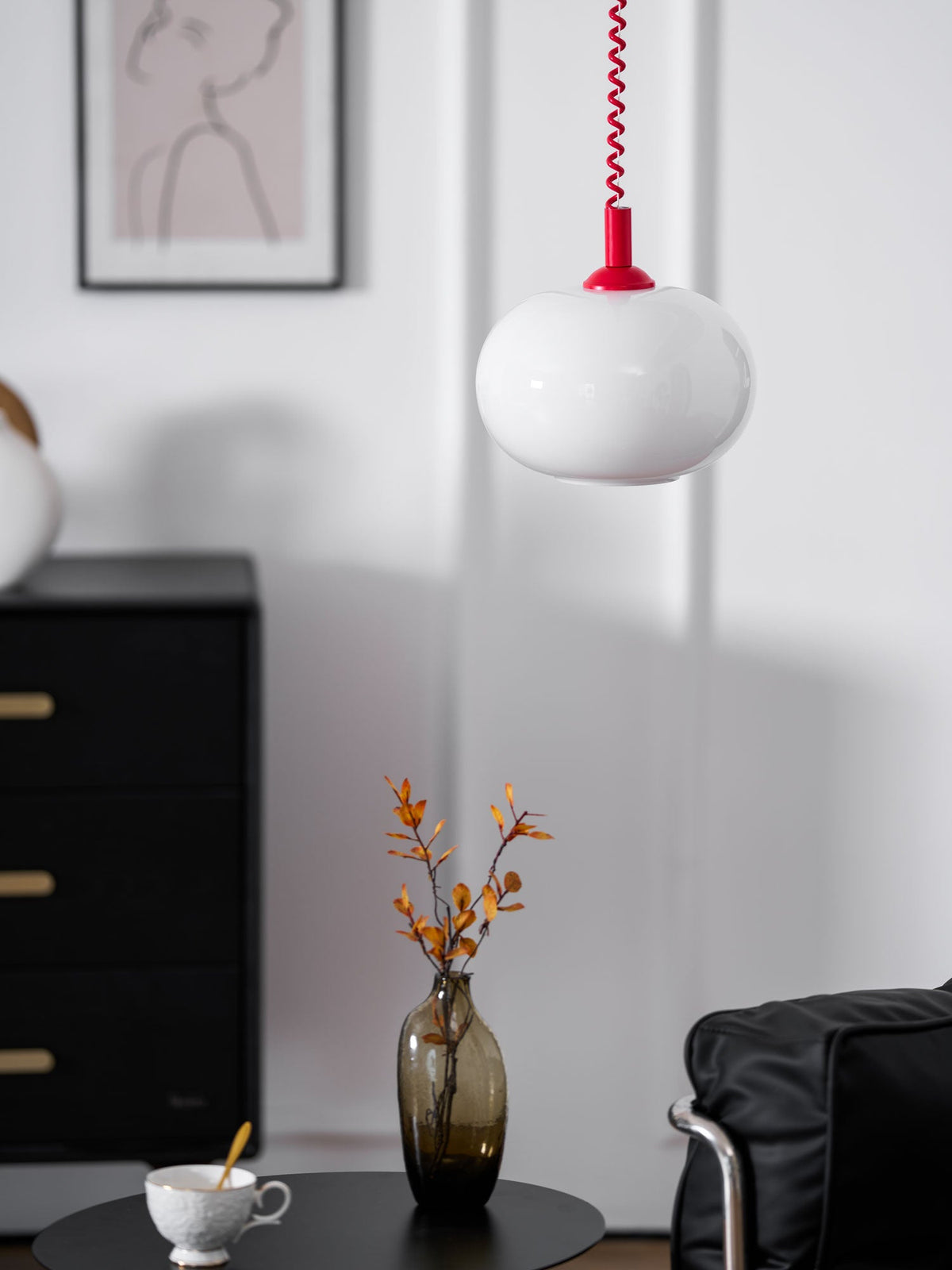 Red-capped Clérachic pendant in front of a framed sketch, a black chest of drawers and a vase of orange leaves.
