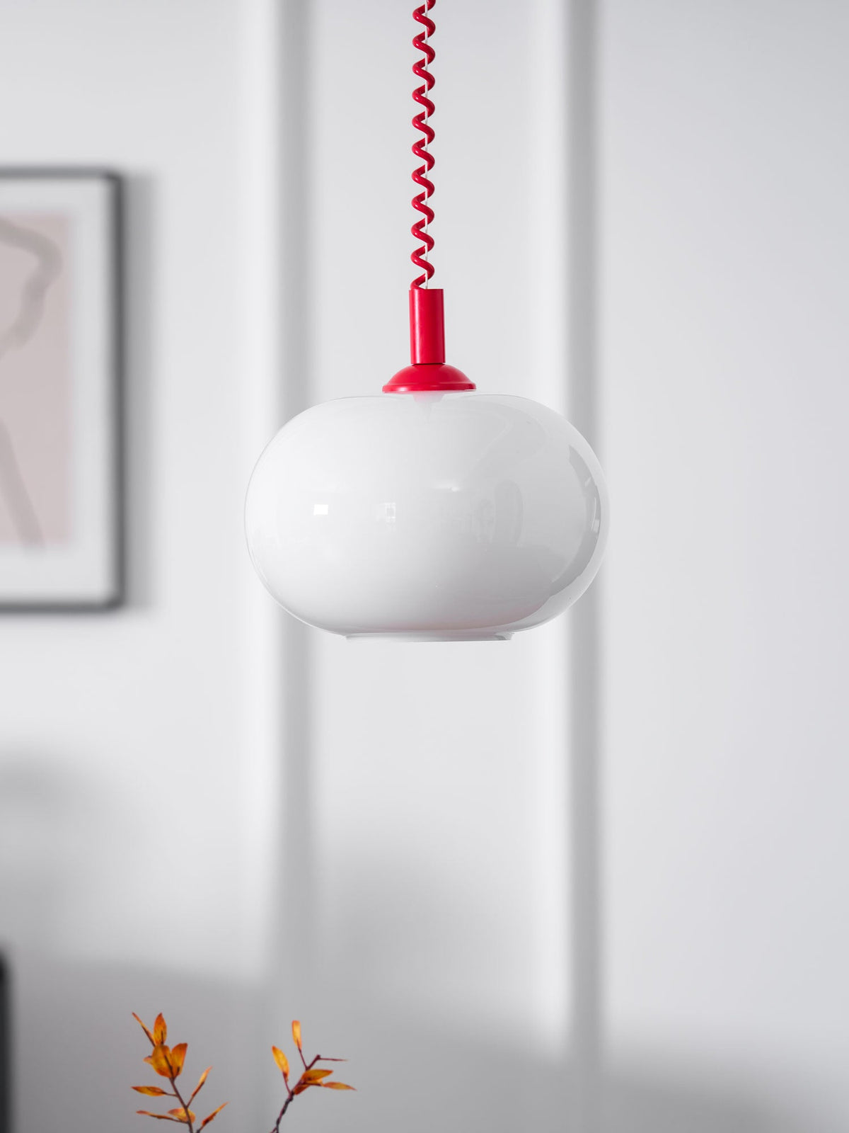 Close view of the white glass shade and red cap of this Clérachic pendant, with a framed picture at the edge of the wall.