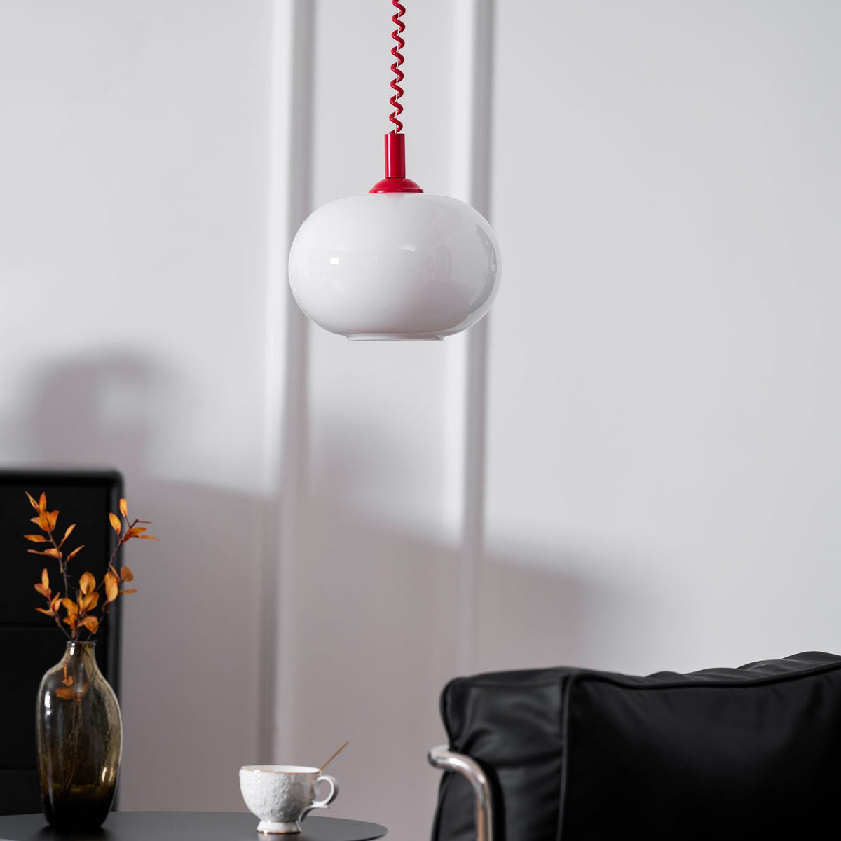 This Clérachic pendant with a red cap hanging beside a black chair and a smoky glass vase with a cup on the table.