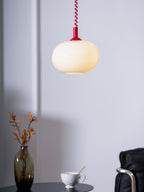 Red-capped Clérachic pendant glowing softly against a lilac-grey wall, with a vase of leaves and a cup below.