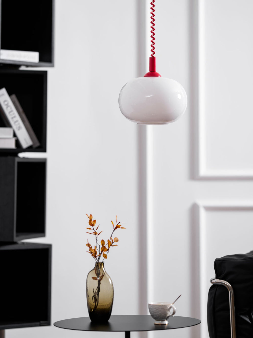 Clérachic pendant with a red cap and white shade hanging by a black shelving unit, a vase of orange leaves below.