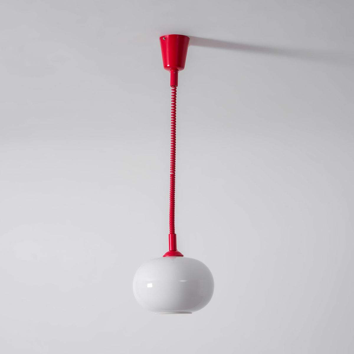 The red Clérachic pendant hanging from a red ceiling cap on a straight red cable, with a white glass shade against a grey wall.