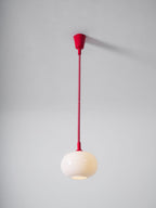 Lit Clérachic pendant hanging from a red ceiling cap on a straight red cable, the cream glass shade glowing softly against grey.