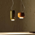 Two Clérachic pendants, one tall olive-green and one orange, glowing against a pale plywood wall above a worktop.