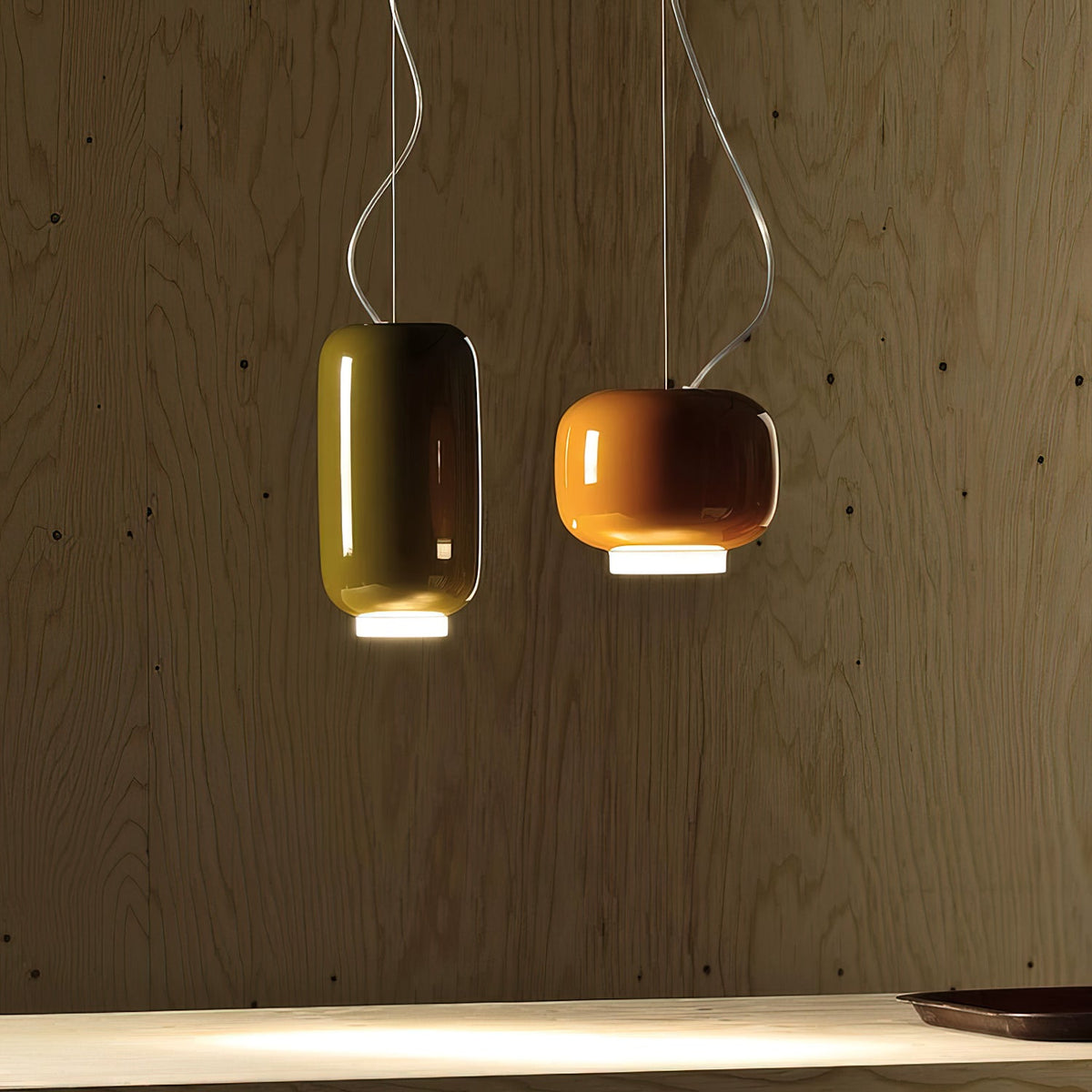 Two Clérachic pendants, one tall olive-green and one orange, glowing against a pale plywood wall above a worktop.