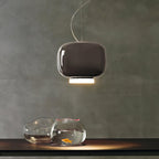 Rounded dark taupe-brown glass shade with a lit white base, a Clérachic pendant above a table with two glass fish bowls.