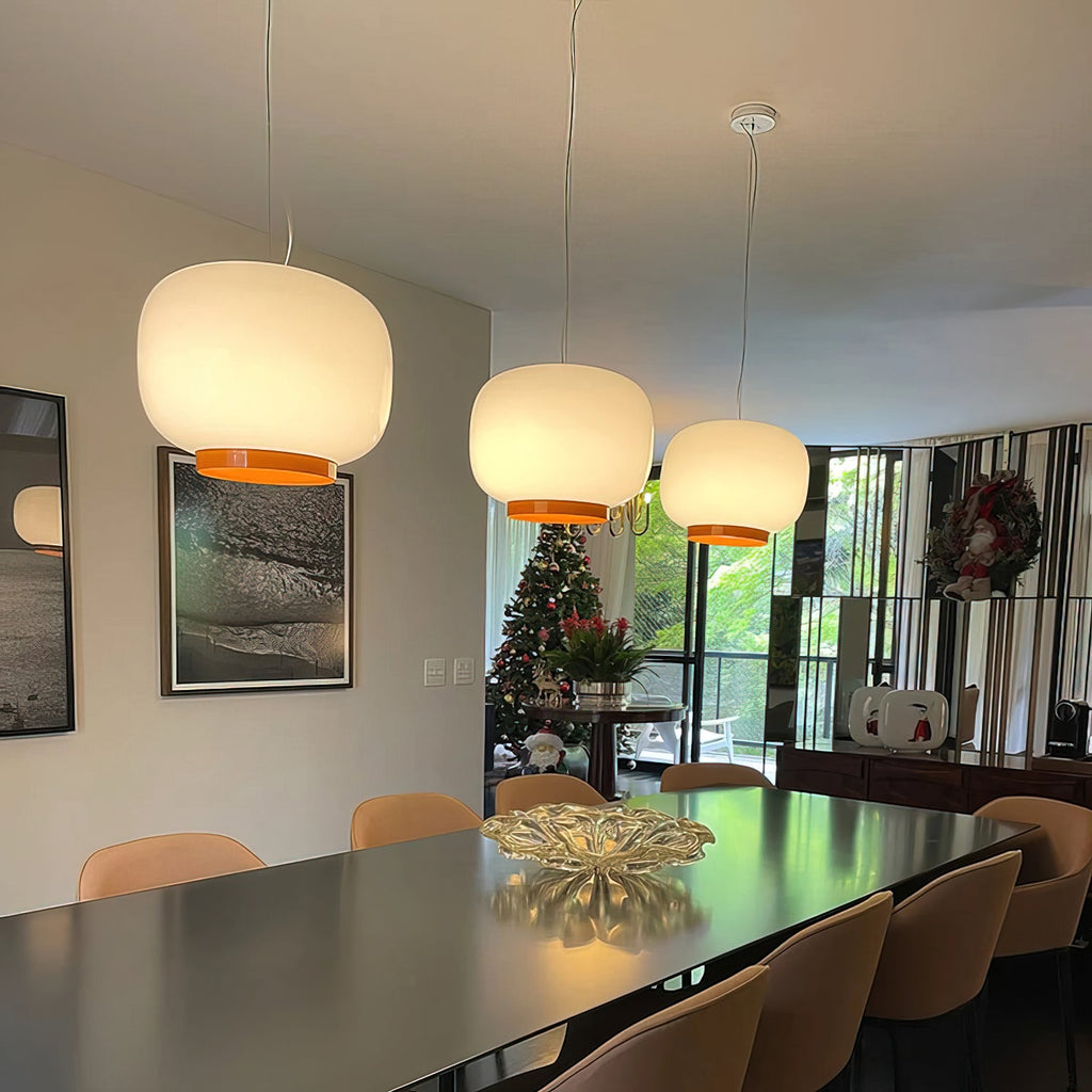 Three white glass pendants with orange bands, Clérachic lanterns glowing over a long dining table in a family room.