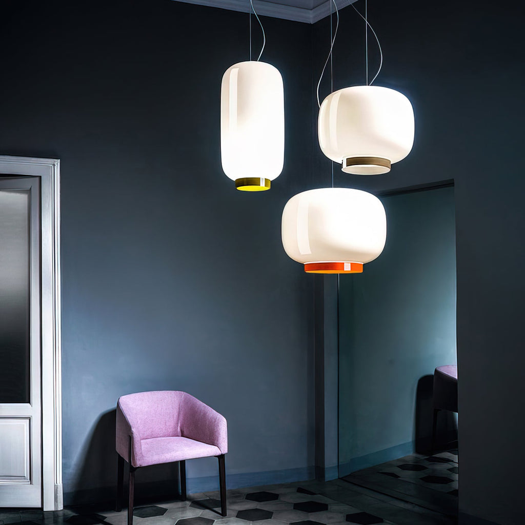 Clérachic pendants in white glass with orange and yellow bands, hung together against a dark blue-green wall beside a pink armchair.