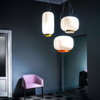Clérachic pendants in white glass with orange and yellow bands, hung together against a dark blue-green wall beside a pink armchair.
