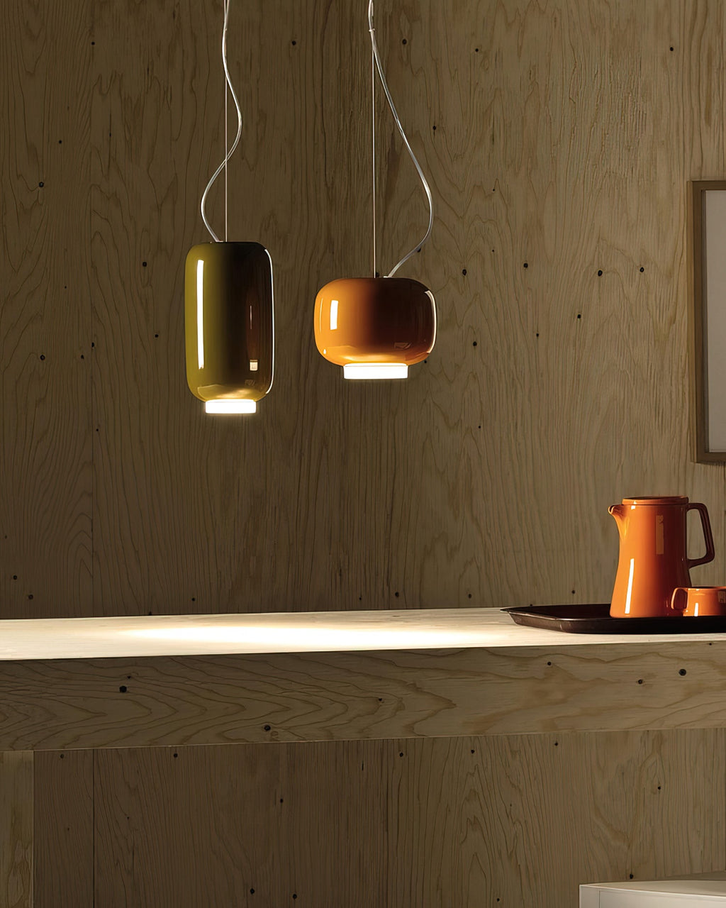 Olive-green and orange glass pendants from Clérachic against a wood-panelled wall, with an orange jug on the shelf below.
