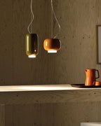 Olive-green and orange glass pendants from Clérachic against a wood-panelled wall, with an orange jug on the shelf below.