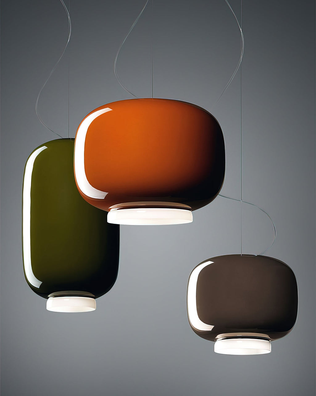 Three glossy glass shades from Clérachic overlapping on a grey backdrop, in orange, green and a dark taupe.