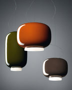 Three glossy glass shades from Clérachic overlapping on a grey backdrop, in orange, green and a dark taupe.