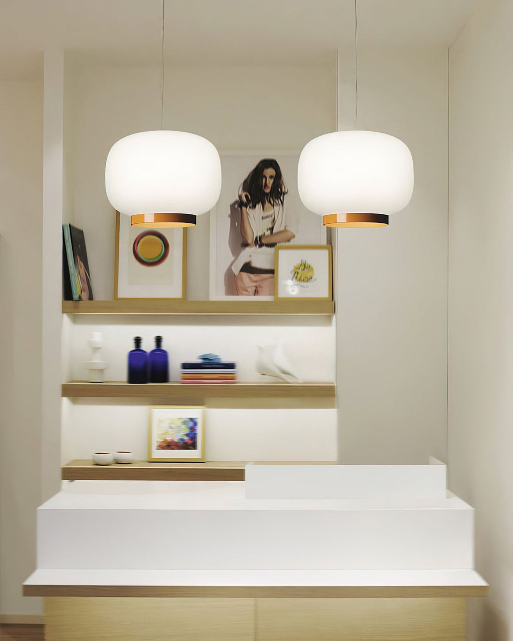 Two white glass lantern pendants with copper-toned bands, Clérachic lights hung over a white counter and open shelves.