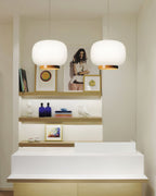 Two white glass lantern pendants with copper-toned bands, Clérachic lights hung over a white counter and open shelves.