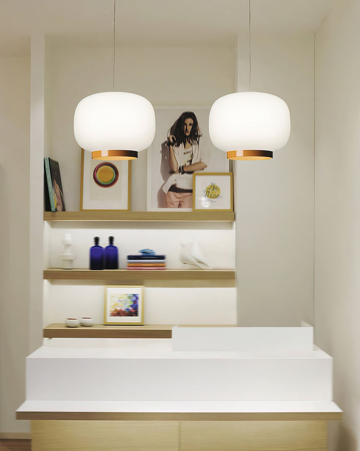 Two white glass lantern pendants with copper-toned bands, Clérachic lights hung over a white counter and open shelves.