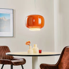 Glossy orange glass pendant above a small round table with cream vases, a Clérachic light beside leather chairs.