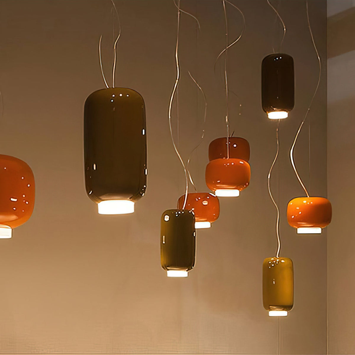 Cluster of Clérachic glass pendants in olive, orange and dark taupe at different heights, hung against a warm wall.