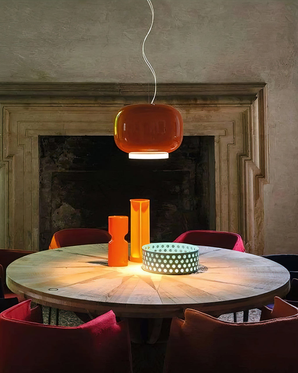 Orange glass pendant from Clérachic above a round wooden table with orange vases, in front of a stone fireplace.