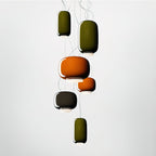 Six Clérachic glass pendants in green, orange and dark tones hang at staggered heights against a pale studio backdrop.