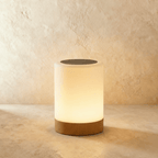 Clérachic's table lamp in a straight cylinder form, its frosted shade glowing warm above a light wood base.