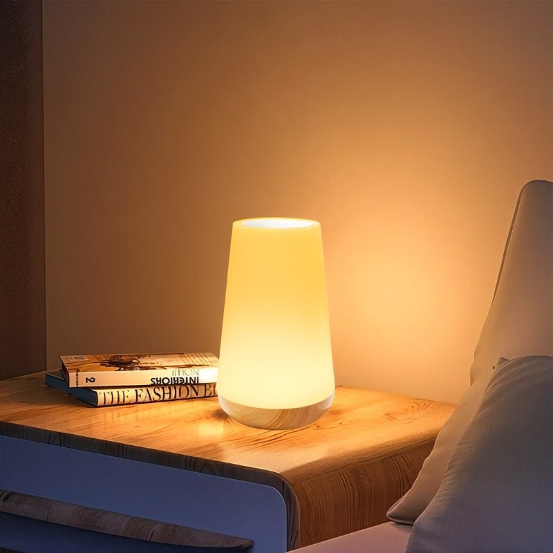 Clérachic's table lamp in its tapered shape, glowing warm on a wood nightstand beside a stack of books.