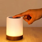 A hand touches the top of the Clérachic table lamp, its frosted cylinder shade glowing warm on a wood base.