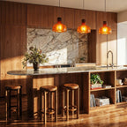 Row of three Clérachic amber glass pendants glowing over a marble kitchen island with walnut cabinets and wooden stools.