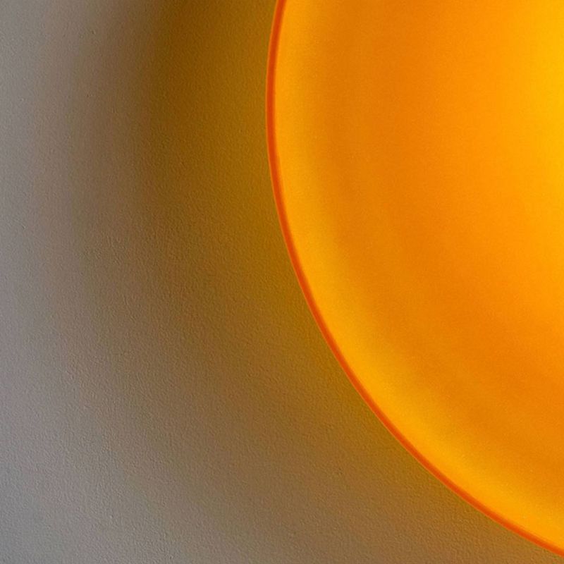 Close-up of the orange glass drum flush mount by Clérachic, lit so the glass glows marigold and washes colour across the wall.