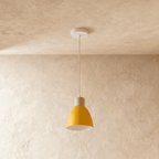 A single Clérachic wood disc pendant light in mustard yellow hangs from a round white ceiling canopy on a plaster wall.