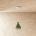 A single Clérachic wood disc pendant light in sage green hangs from a round ceiling canopy over a plaster wall.