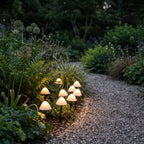 A cluster of Clérachic solar mushroom stake lights glows warm white beside a gravel path in a shaded evening garden bed.