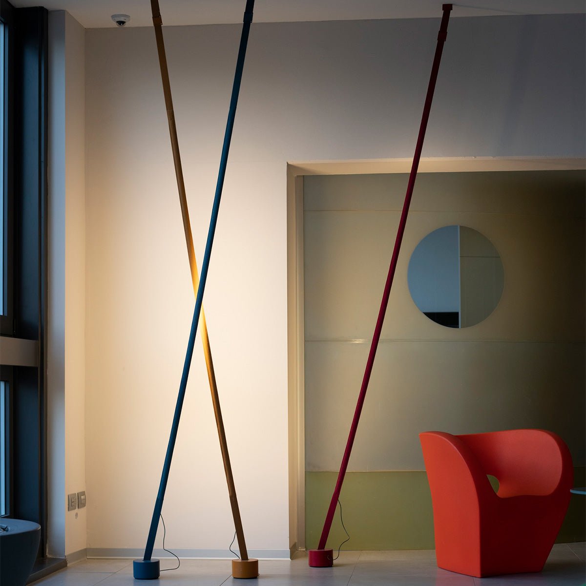 Three Clérachic floor lamps in gold, blue and red cross each other at steep angles in a bright room with an orange armchair.