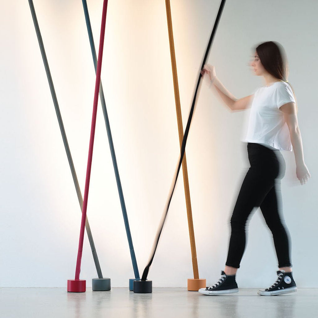 A woman walks past a row of leaning Clérachic floor lamps in red, grey, blue, ochre and black against a softly lit white wall.