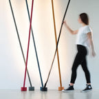 A woman walks past a row of leaning Clérachic floor lamps in red, grey, blue, ochre and black against a softly lit white wall.