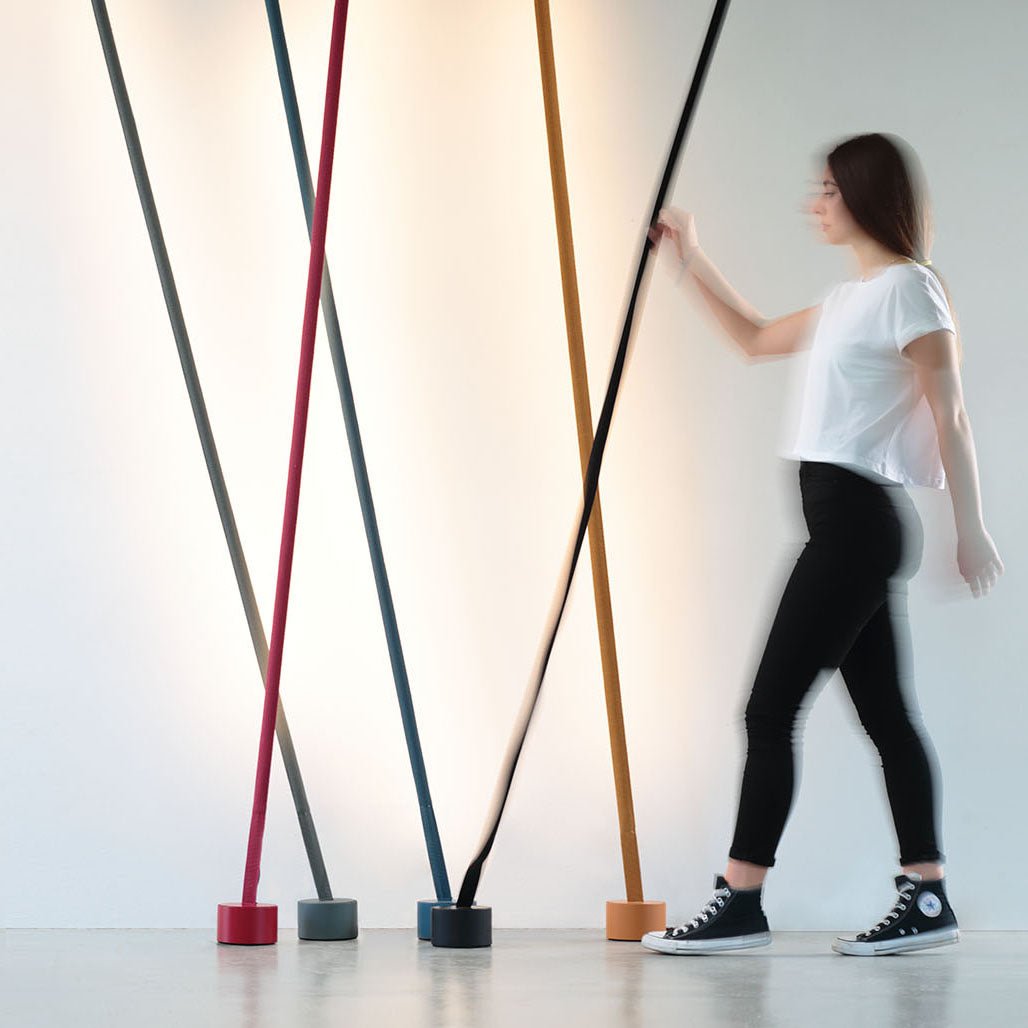 A woman walks past a row of leaning Clérachic floor lamps in red, grey, blue, ochre and black against a softly lit white wall.
