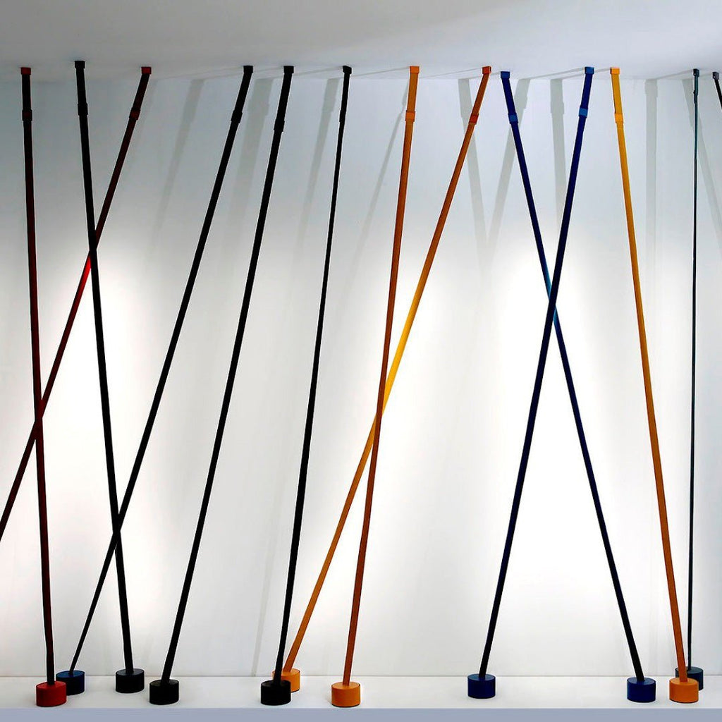 A row of Clérachic floor lamp strips in black, blue, ochre and red lean at crossing angles against a white wall, each on a round base.
