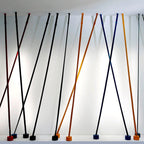 A row of Clérachic floor lamp strips in black, blue, ochre and red lean at crossing angles against a white wall, each on a round base.