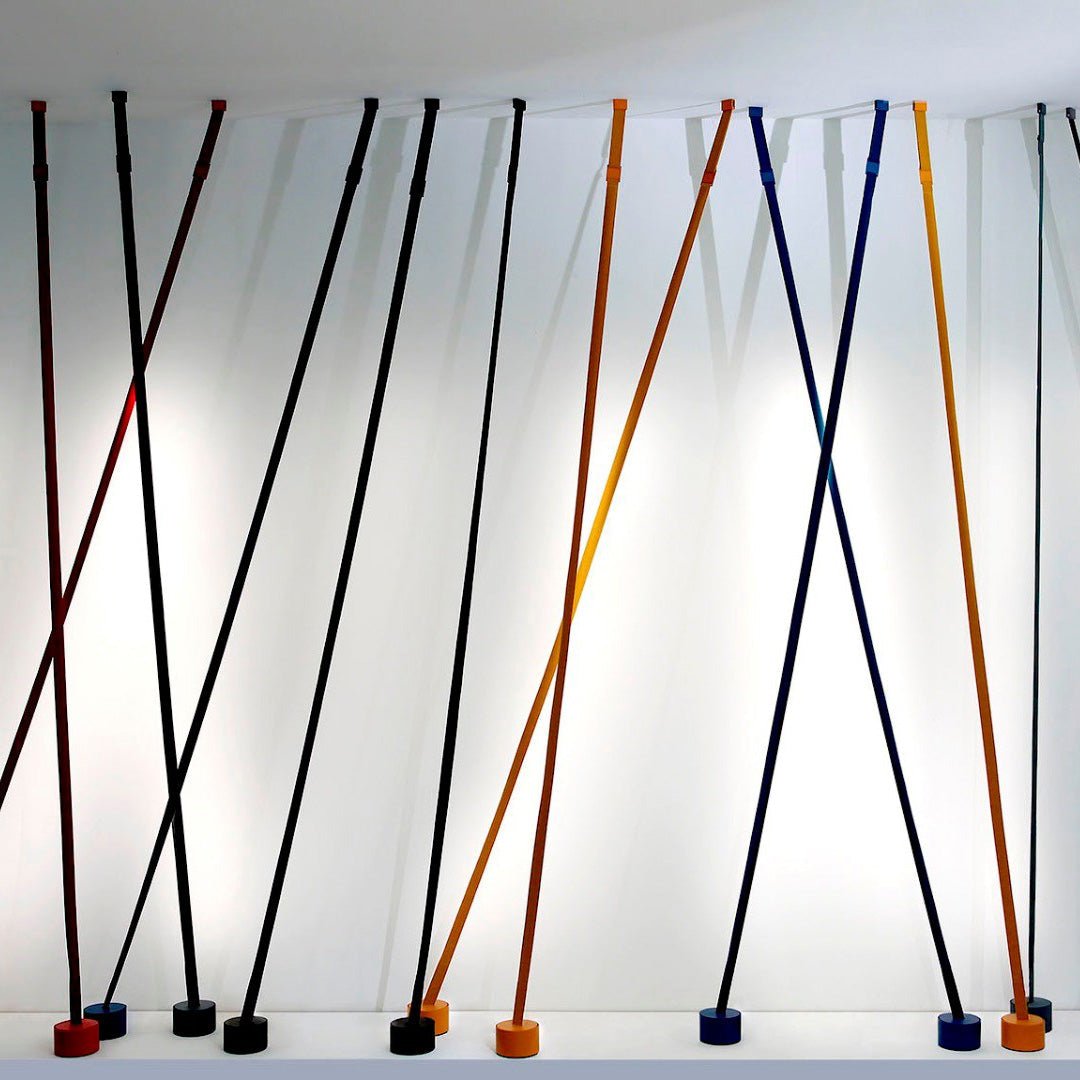 A row of Clérachic floor lamp strips in black, blue, ochre and red lean at crossing angles against a white wall, each on a round base.