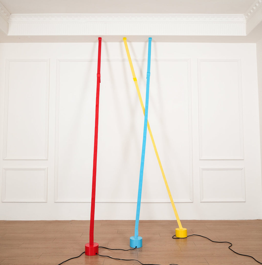 Three Clérachic floor lamp strips in red, light blue and yellow rise from round bases on a wood floor to a plain ceiling moulding.