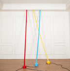 Three Clérachic floor lamp strips in red, light blue and yellow rise from round bases on a wood floor to a plain ceiling moulding.