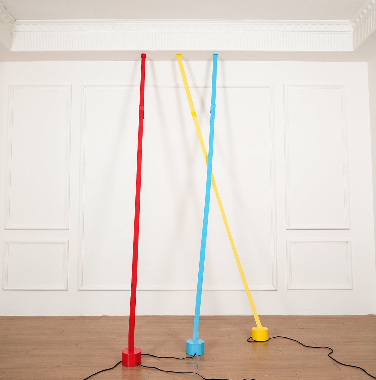 Three Clérachic floor lamp strips in red, light blue and yellow rise from round bases on a wood floor to a plain ceiling moulding.