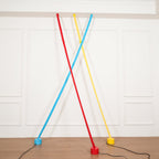 A red and a light blue Clérachic floor lamp strip cross in front of a yellow one, all on round bases above a wood floor.