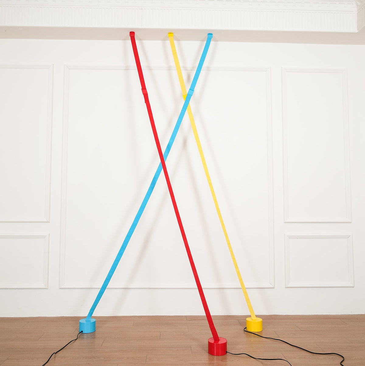 A red and a light blue Clérachic floor lamp strip cross in front of a yellow one, all on round bases above a wood floor.