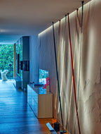 Seen along a plaster wall in a long living room, several Clérachic floor lamp strips run up to a lit ceiling edge by a window.