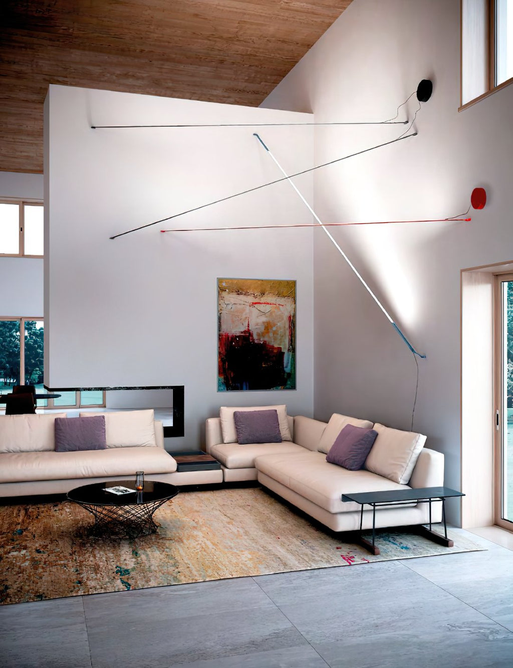 Thin lines of light from Clérachic floor lamps stretch across a high white wall in a living room with cream sofas and a rug.