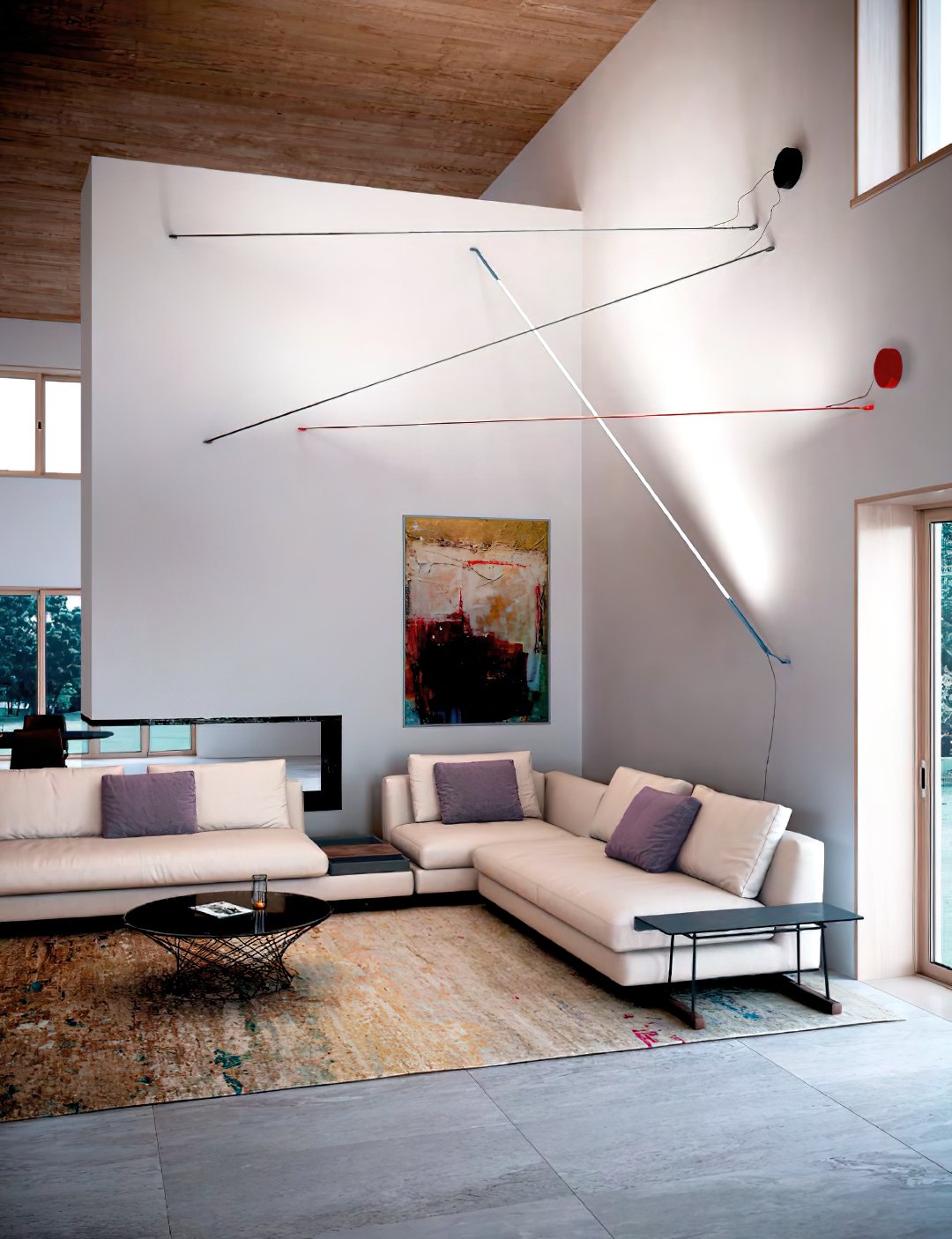 Thin lines of light from Clérachic floor lamps stretch across a high white wall in a living room with cream sofas and a rug.