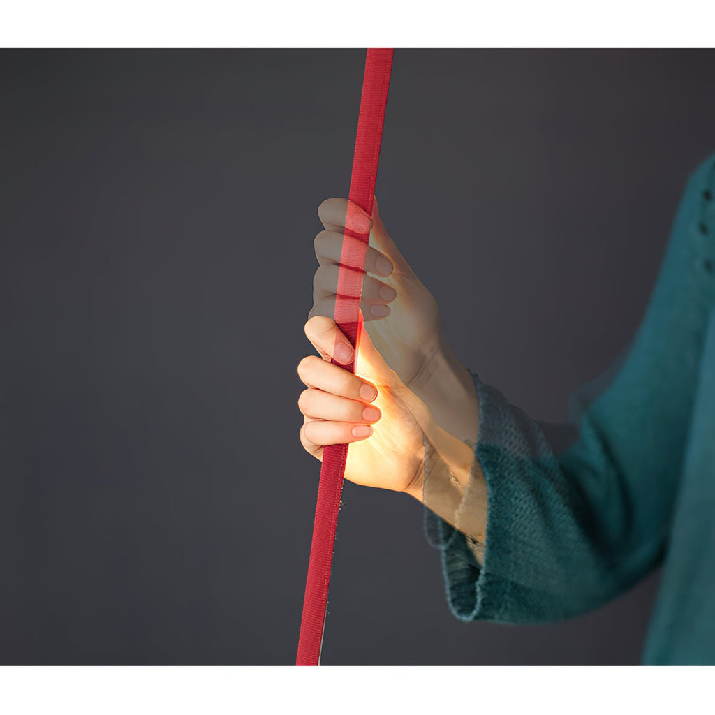 A hand grips a red Clérachic floor lamp strip against a dark grey backdrop, showing how slim and flat the webbing is.