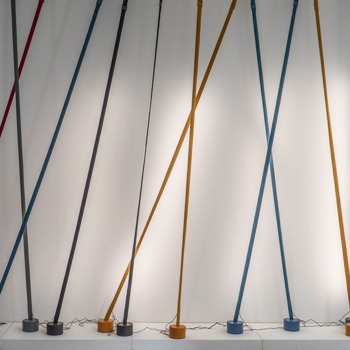 Blue, grey, ochre and black Clérachic floor lamp strips lean against a white wall lit from behind, each on its own round base.